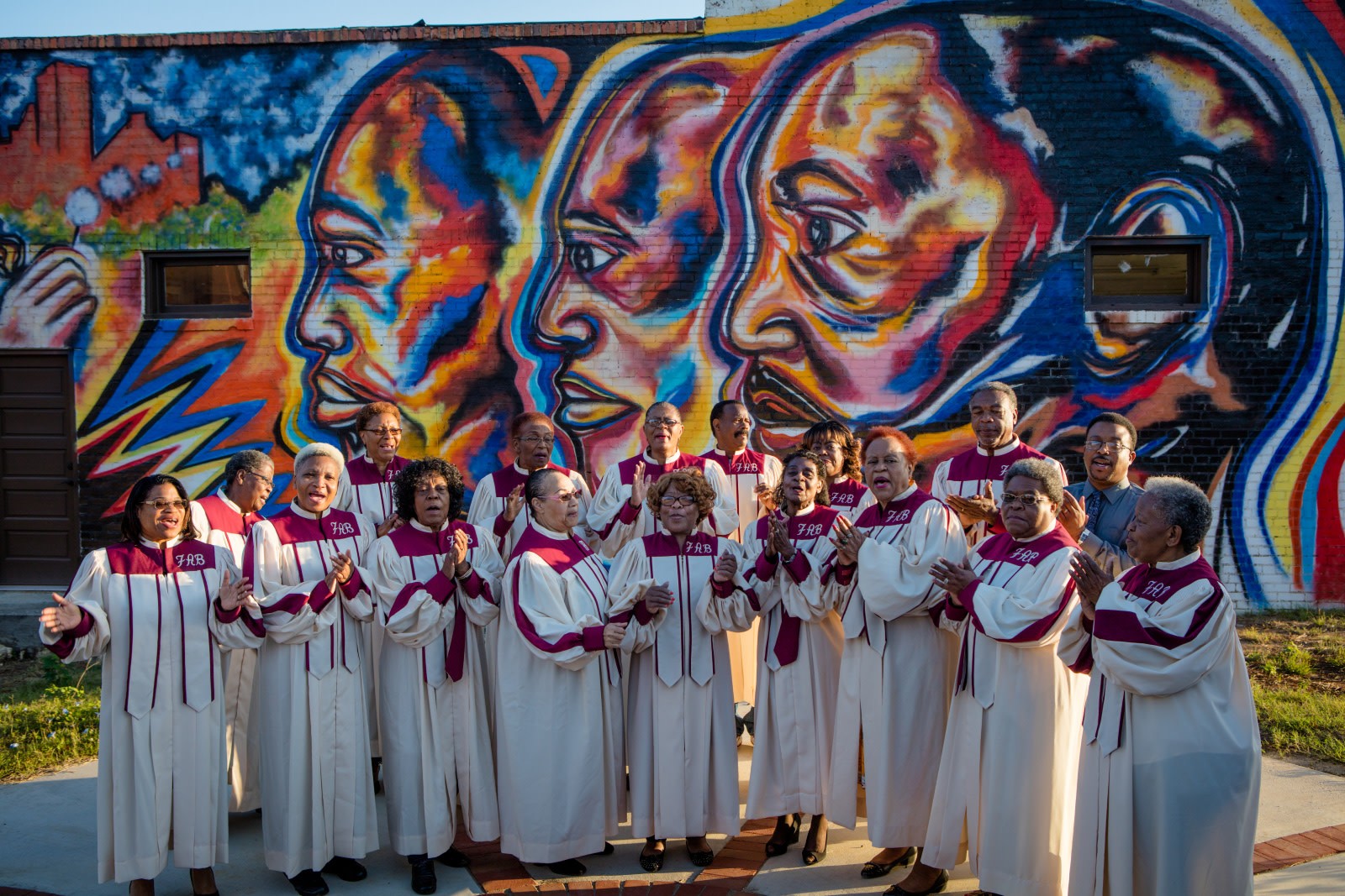 Church choir in robes singing in front of a community mural of three large faces by Corey Barksdale
