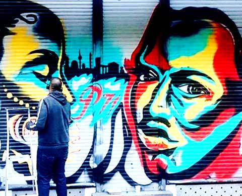 Artist spray painting two large faces and a skyline onto a metal roll-up gate