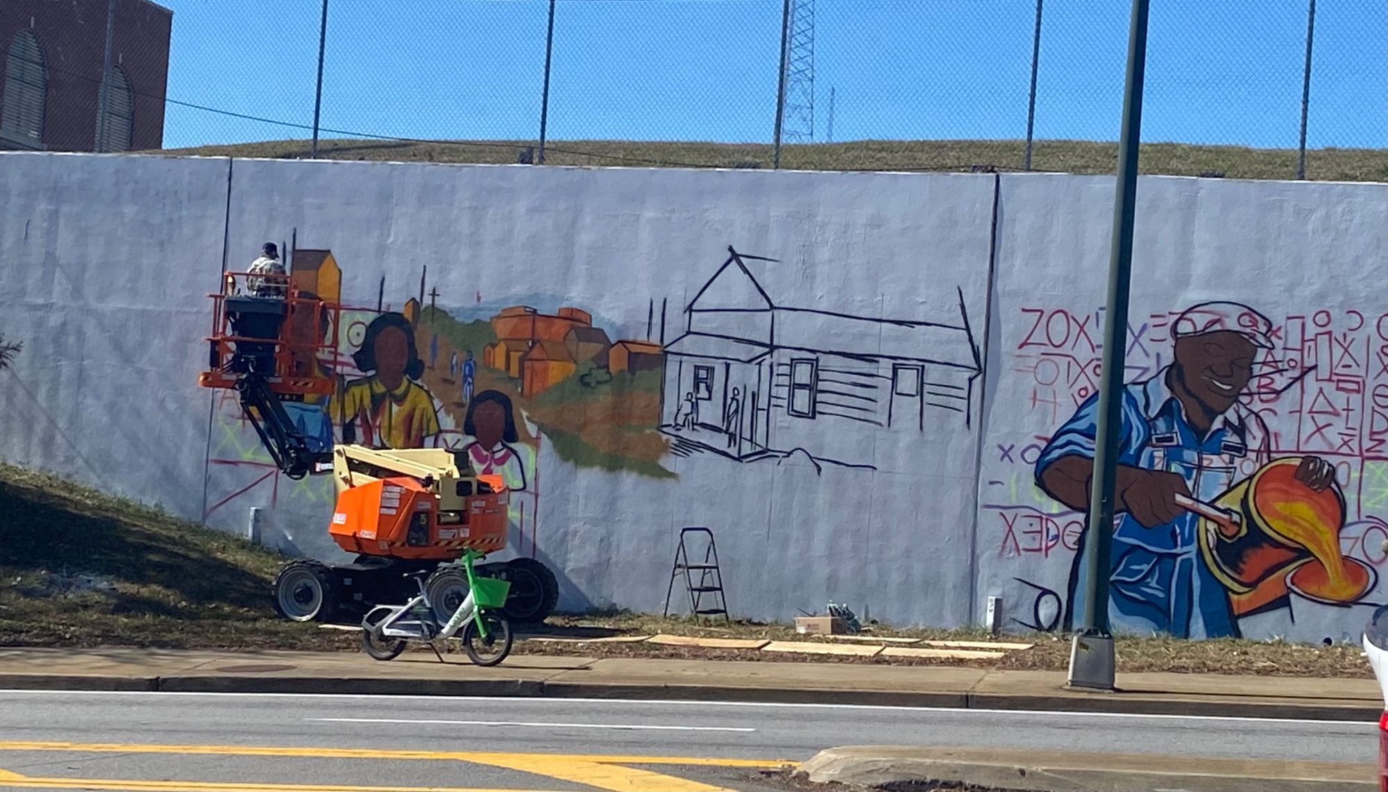 Wide street view of the Blandtown mural wall in progress, boom lift and house outline, Atlanta mural painter Corey Barksdale