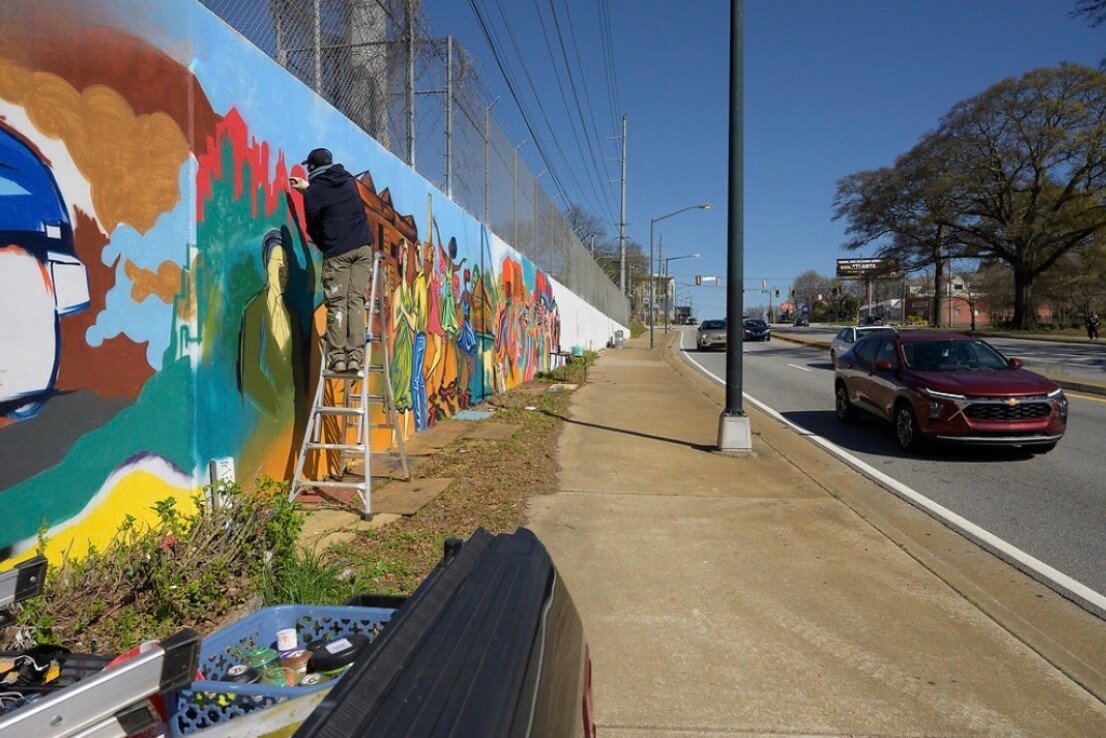Wide view of the Blandtown mural wall at 17th Street with Corey Barksdale painting from a ladder as traffic passes