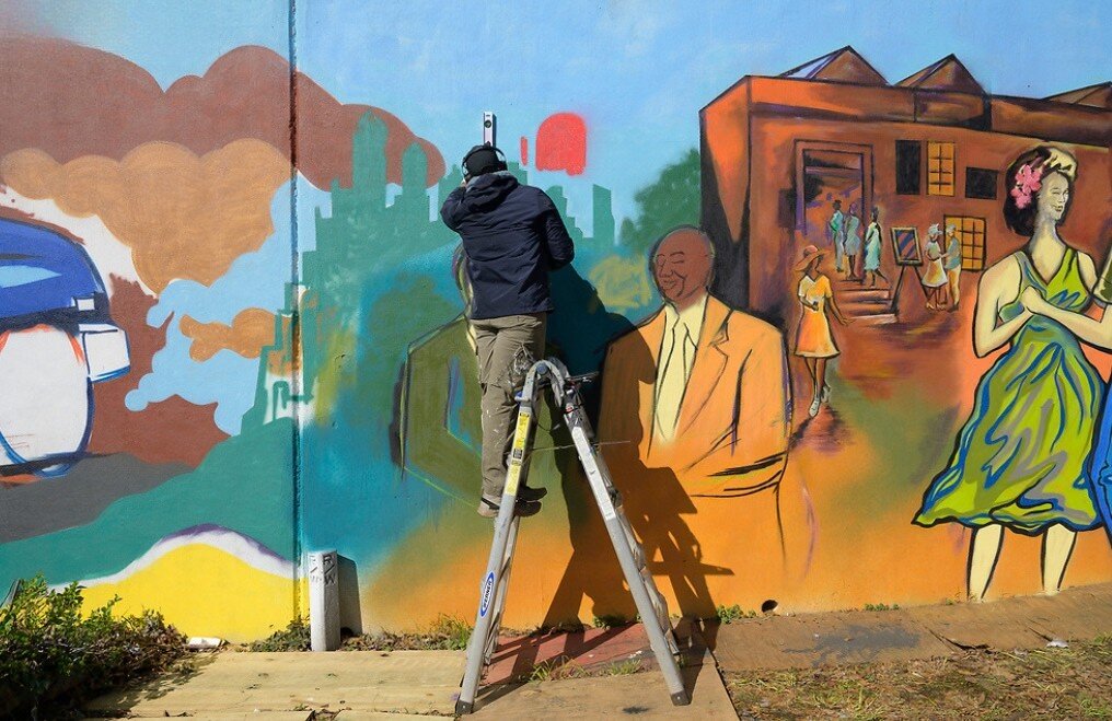Corey Barksdale on a ladder painting a portrait scene beside the barbershop panel of the Blandtown mural