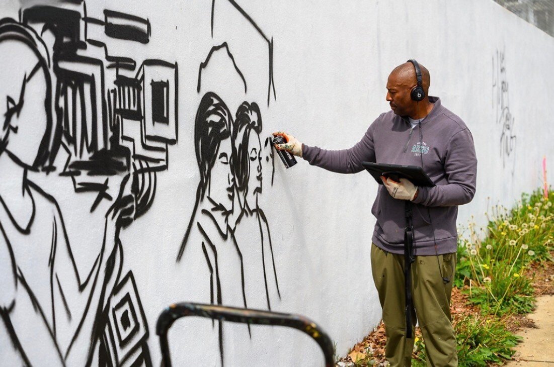 Corey Barksdale freehand-sketching the black outline stage of the Blandtown mural while referencing a tablet